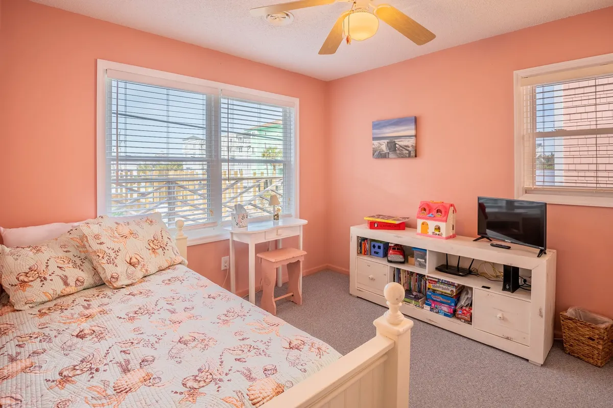 A playful bedroom with a bed topped by a floral quilt, a desk and chair set up by the window, and a television unit with toys, creating a space suitable for kids or young guests to relax, read, and play.