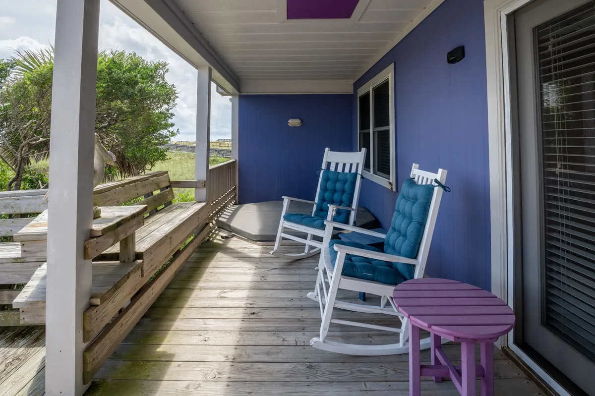 Two classic rocking chairs and a purple accent table sit on a wooden deck with railings, facing out toward open views. The tranquil setting invites moments of relaxation, conversation, and enjoying the outdoors, perhaps while listening to music or awaiting fireworks from the boardwalk.