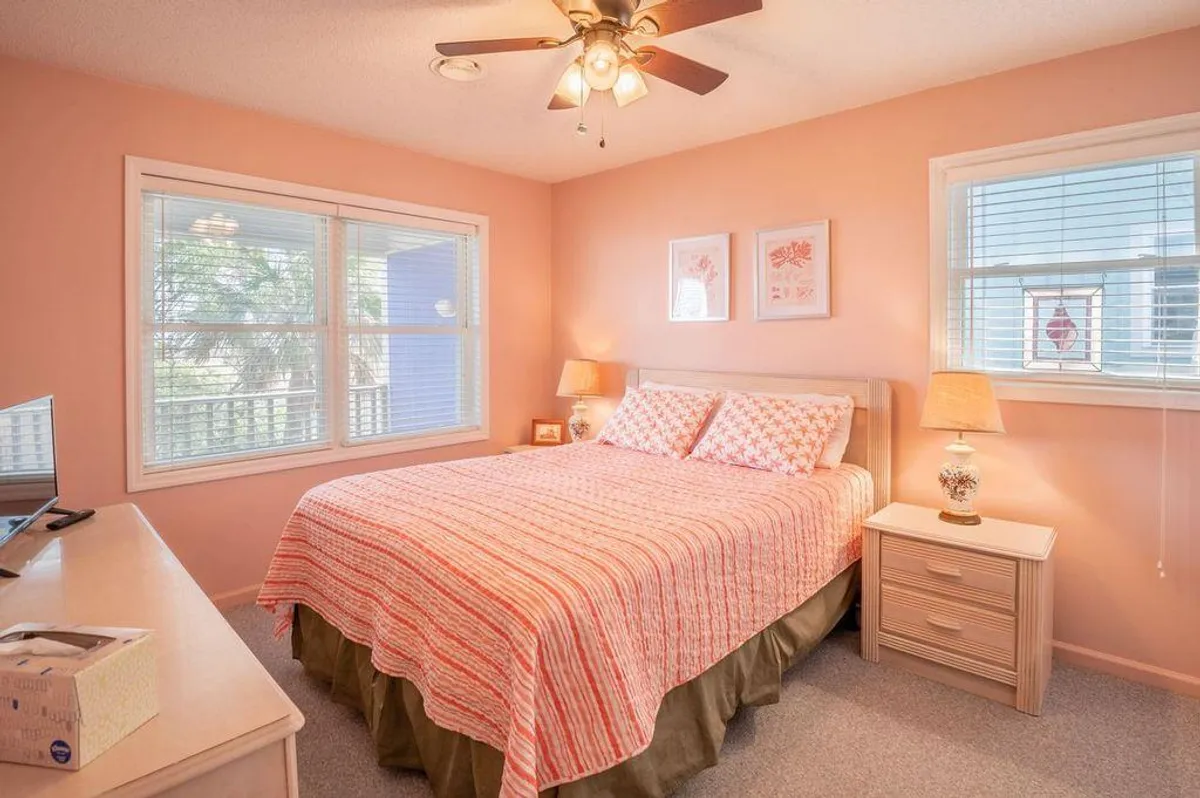 A serene bedroom with a bed featuring patterned bedding, large windows that fill the space with natural light, and a nightstand with a lamp. The overall atmosphere is calm and inviting, perfect for relaxing after a day at the beach.