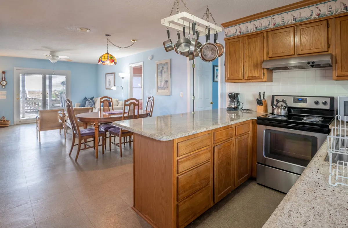 A warm, open kitchen with a large island sits at the center, accented by hanging pots, wooden cabinetry, and a view into the adjacent dining area. The layout feels inviting and functional, ideal for family meal prep or casual breakfasts.