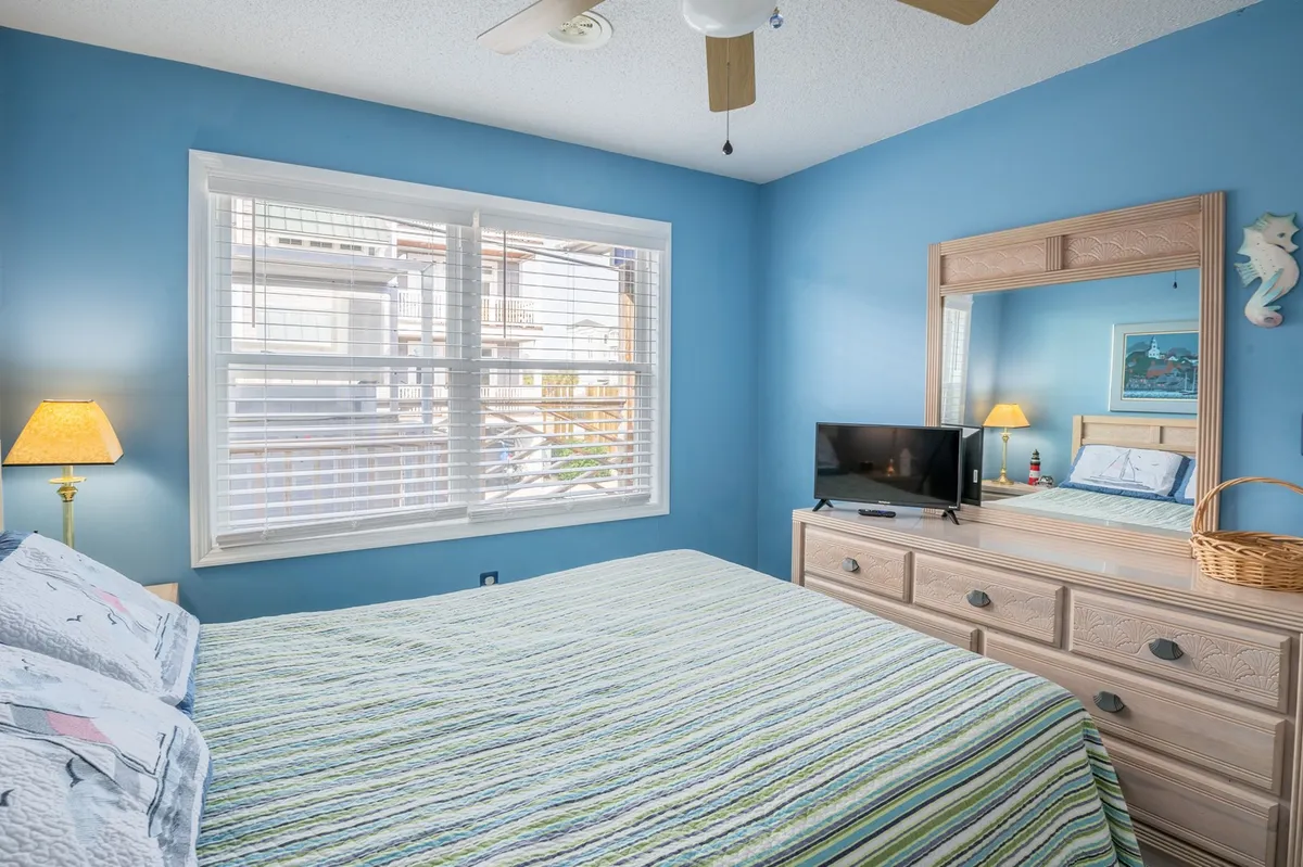 A cozy bedroom with a striped comforter, nightstand with lamp, television on a dresser, and a mirror reflecting the room. Sunlight streams in from the window, completing a restful and homey vibe.