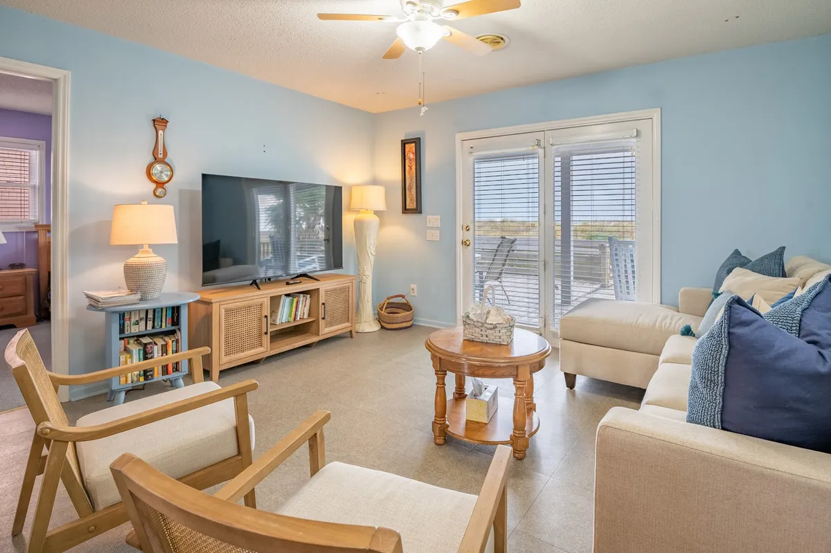 A cozy living room featuring a sofa with accent pillows, a coffee table, a large television, and sliding glass doors that let in bright natural light. The setup is ideal for relaxing, socializing, and family entertainment.