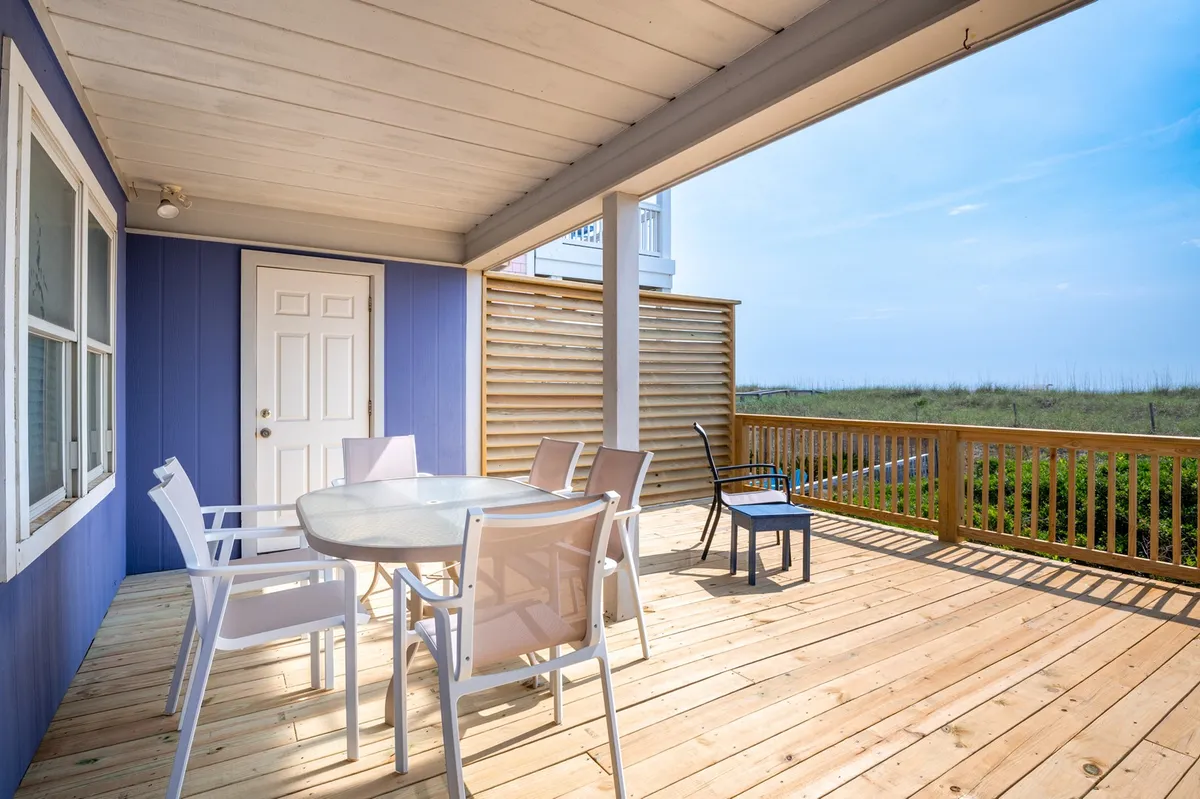 This exterior image features a patio dining table and chairs set against a backdrop of natural landscape, with a door invitingly open to the outside. The view evokes the charm of al fresco living and the sense of connection to the beach and surrounding area.