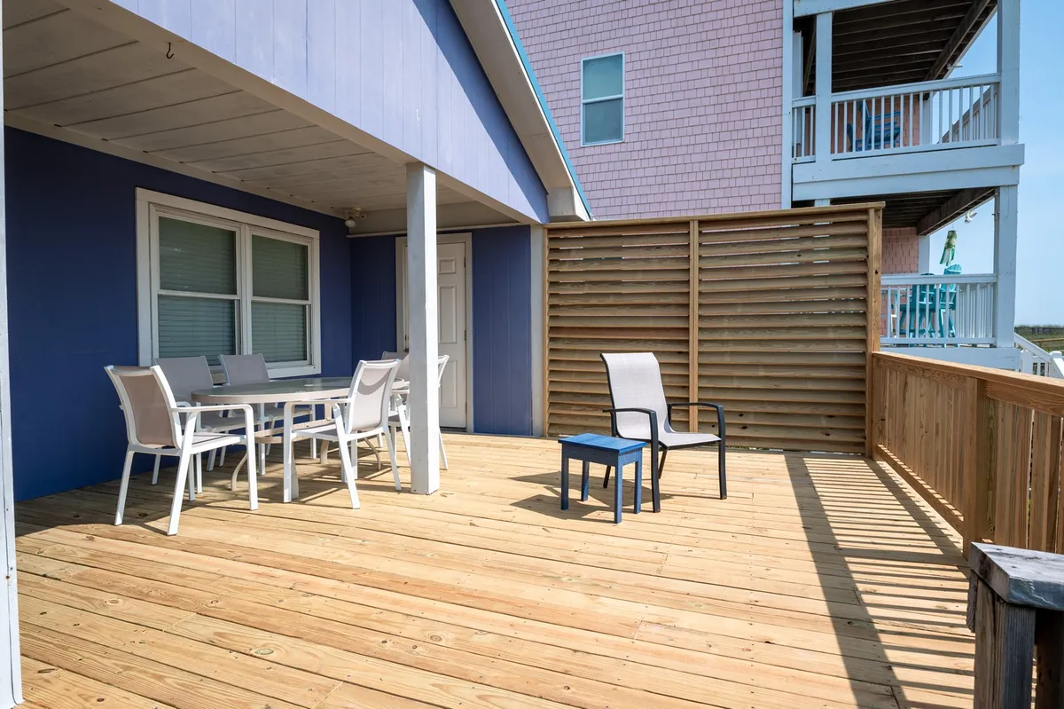 This image features a wooden deck furnished with a table and chairs set against a blue wall, providing a shaded, casual atmosphere ideal for relaxing or dining outdoors. The setting suggests easy access from the home to the outdoors, with a seamless blend between indoor comfort and the inviting beach environment.