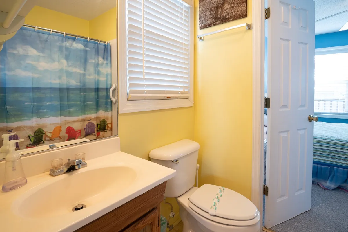 A cheerful bathroom featuring a vanity with a sink, toilet, sunny yellow walls, and a shower curtain with a playful beach scene. The vibe is light-hearted and refreshing, ideal for unwinding or starting the day.