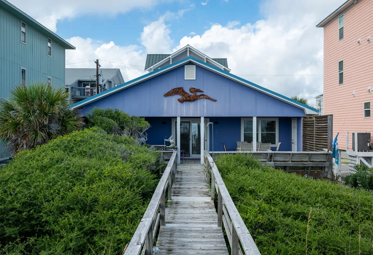 This photo highlights the blue house exterior, a wooden walkway leading to the entrance, lush palm trees, and a decorative eagle sculpture. The inviting approach showcases the covered area and hints at protected, practical parking beneath the structure.