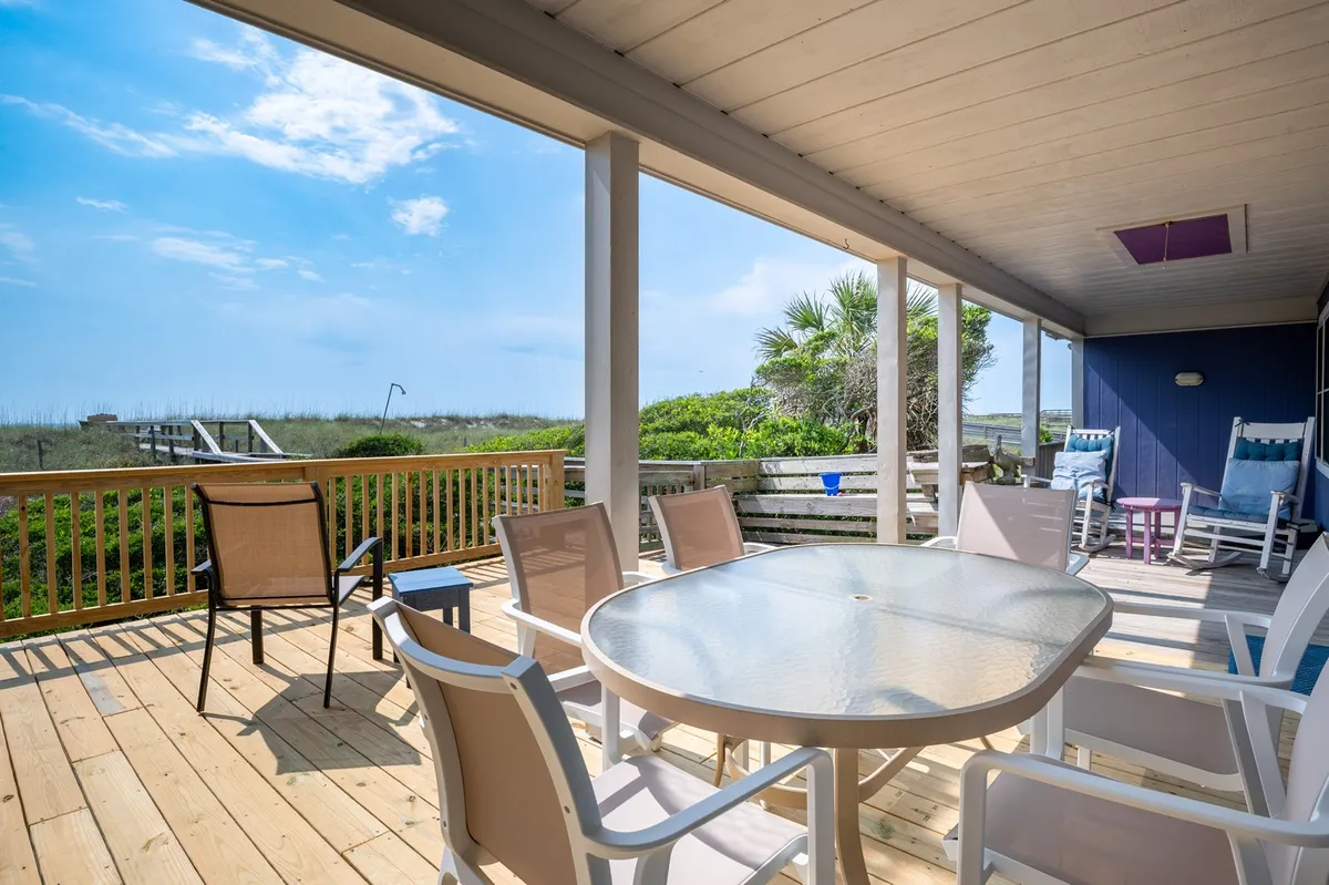 A wide view of the porch featuring a dining table with chairs, several inviting rocking chairs, and lush greenery in the background. The porch looks out onto the beach, blending indoor comfort with panoramic ocean views.