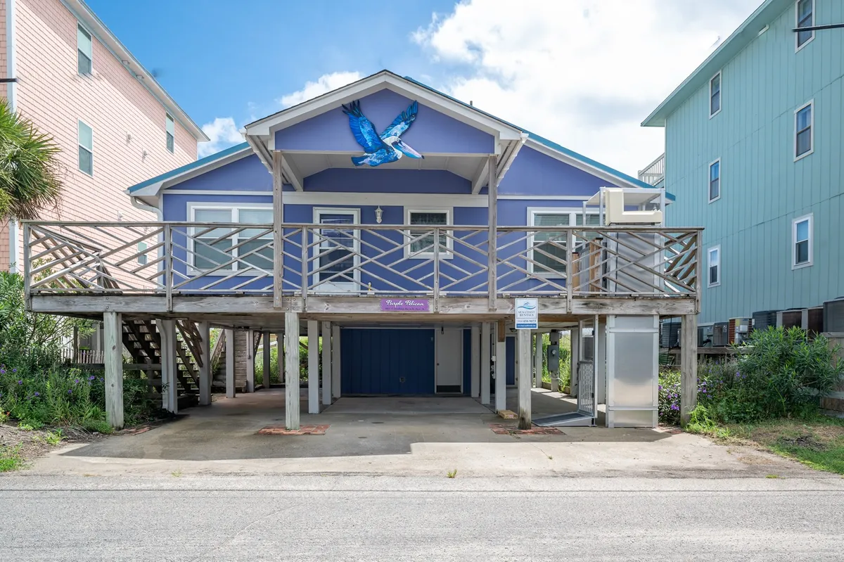 The image shows the vibrant purple exterior of the Purple Pelican beach house, featuring a striking blue bird mural and a spacious wooden porch overlooking the ocean. The scene evokes a cheerful, laid-back atmosphere, with hints of relaxation and connection, ideal for families or friends looking to unwind by the beach.