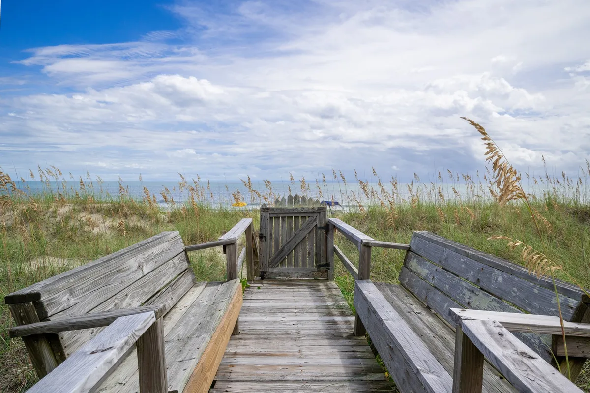 A scenic angle showing wooden benches thoughtfully placed on a raised platform overlooking the ocean. An open gate leads out to stunning sea vistas, inviting moments of reflection or group gatherings to watch sunsets or fireworks.