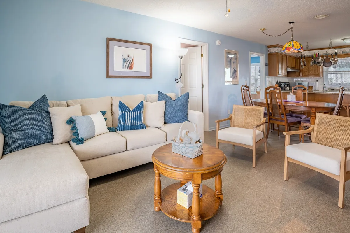 A comfortable living area with a sofa adorned with decorative cushions, a round wooden coffee table, and a dining area visible in the background. The space feels open and encourages relaxation and gathering, with clear pathways for easy movement.