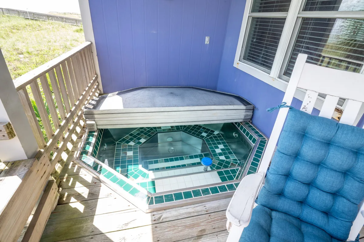 A tranquil scene on the wooden deck displaying a sunken hot tub, a comfortable rocking chair, and sweeping views toward the horizon. The space radiates serenity, perfect for soaking and unwinding after a day at the beach.