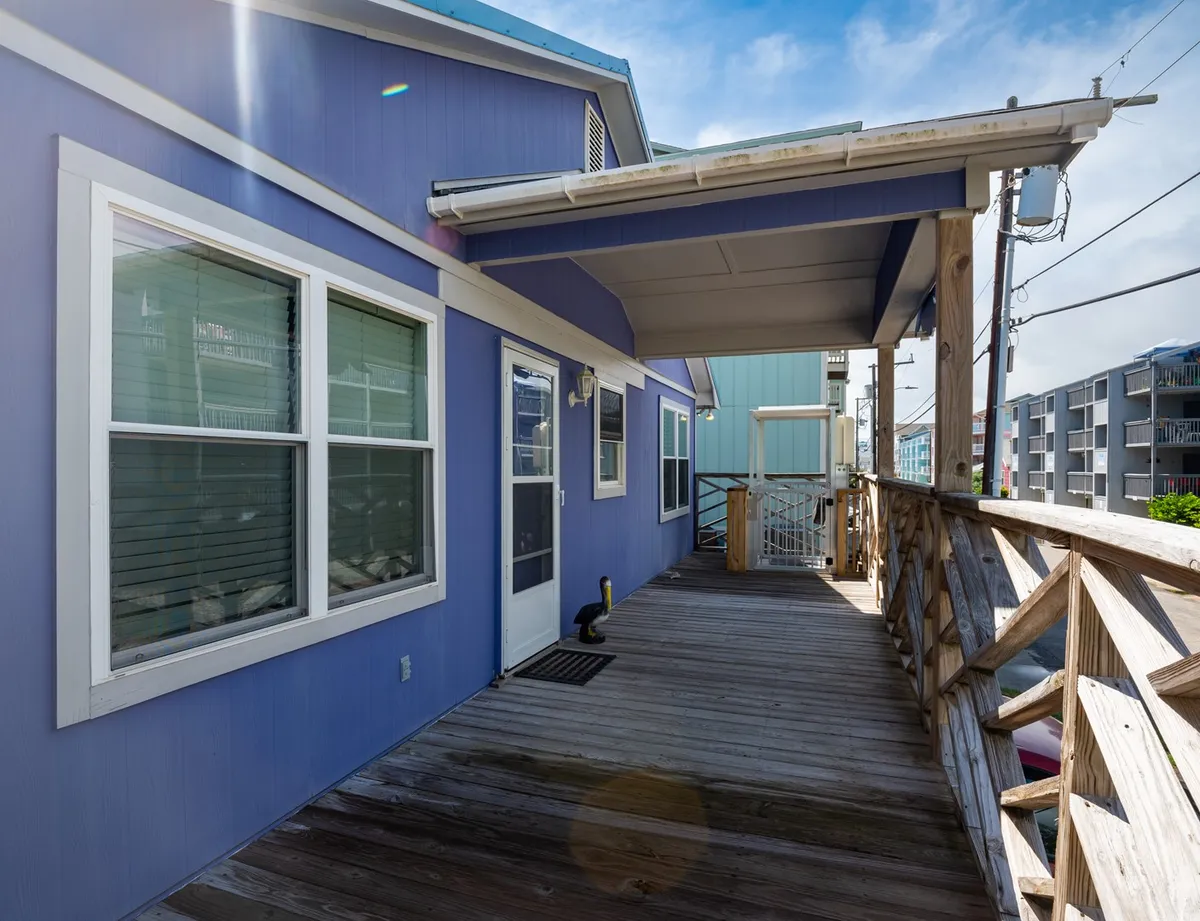The image presents a blue exterior with wooden decking and a doorway, inviting guests into or out of the house. The deck is designed for relaxing and entertaining, with plenty of space and a sense of privacy, making it ideal for unwinding after time on the beach.
