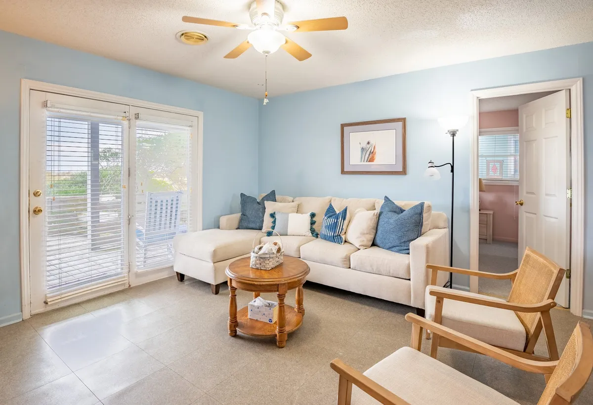 A cozy living room with a sofa adorned with cushions, a coffee table, and double doors leading outside. Artwork on the wall and soft lighting create an inviting, nostalgic beach-cottage atmosphere.