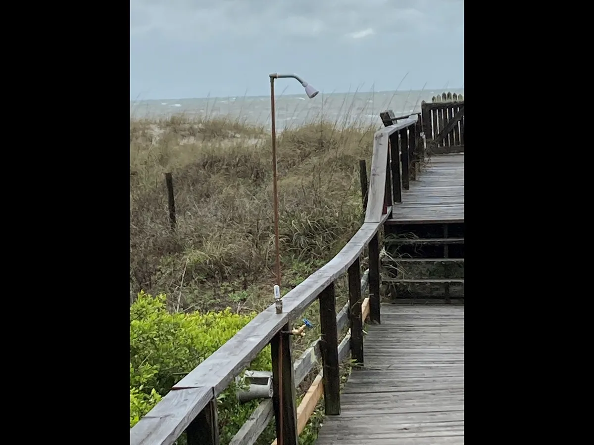 A wooden walkway meanders through beach grass, with glimpses of the ocean in the background and a lamp post adding a nostalgic touch. The scene suggests peaceful family strolls, beautiful sunset views, and a sense of community gathering at the coast.